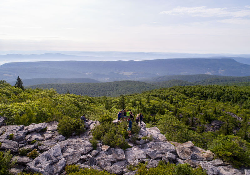 The image captures a scenic view from a rocky outcrop, possibly a mountain peak, overlooking a vast expanse of forested hills and valleys. A group of people is gathered on the rocks, enjoying the view. The sky is overcast, creating soft lighting across the landscape. The overall impression is one of natural beauty and recreational activity.